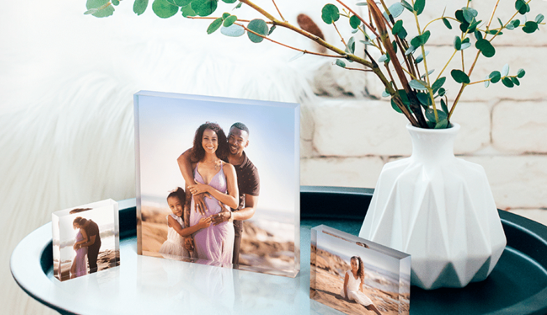 A round table with three acrylic photo blocks displaying family portraits next to a white geometric vase holding leafy branches, set in a bright, modern room.
