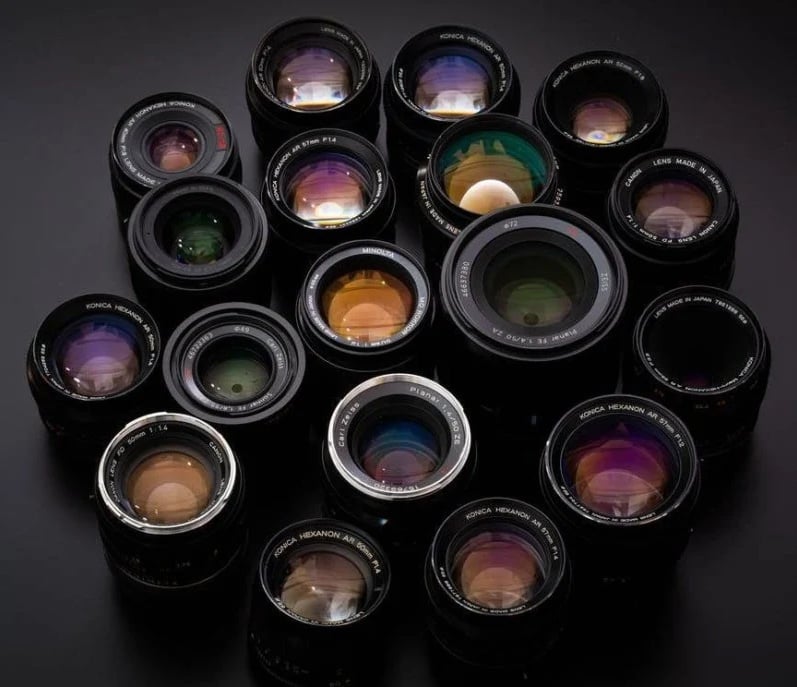 A collection of various camera lenses is arranged closely together on a dark surface, showcasing different sizes and brands with colorful lens reflections visible on the glass elements.
