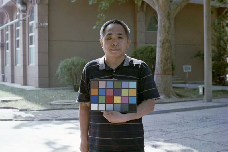 A man stands outdoors on a sunny day holding a color calibration chart. He wears a dark striped polo shirt, and there is a large tree and building in the background.