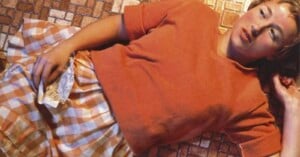 A woman in an orange top and checkered skirt lies on a tiled floor, holding a crumpled note in one hand and looking off to the side with a contemplative expression.