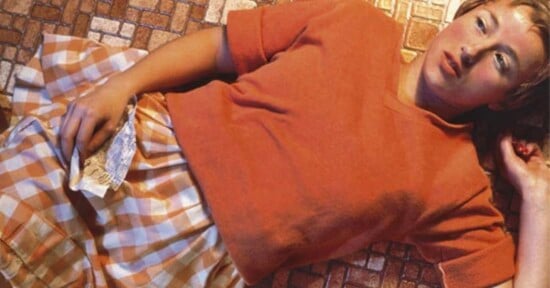 A woman in an orange top and checkered skirt lies on a tiled floor, holding a crumpled note in one hand and looking off to the side with a contemplative expression.