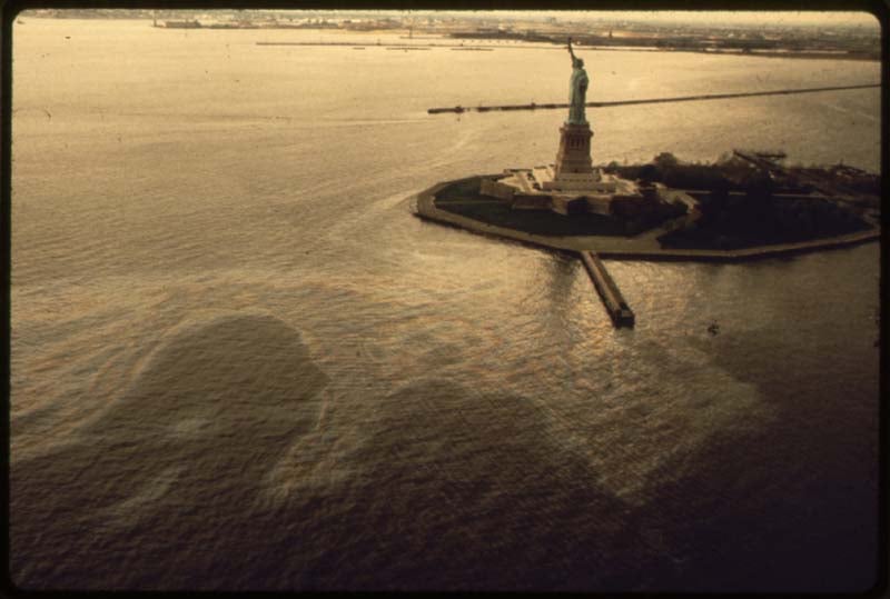 Aerial view of the Statue of Liberty on Liberty Island, surrounded by water with visible ripples and reflections. The horizon and shoreline are visible in the distance under a cloudy sky.