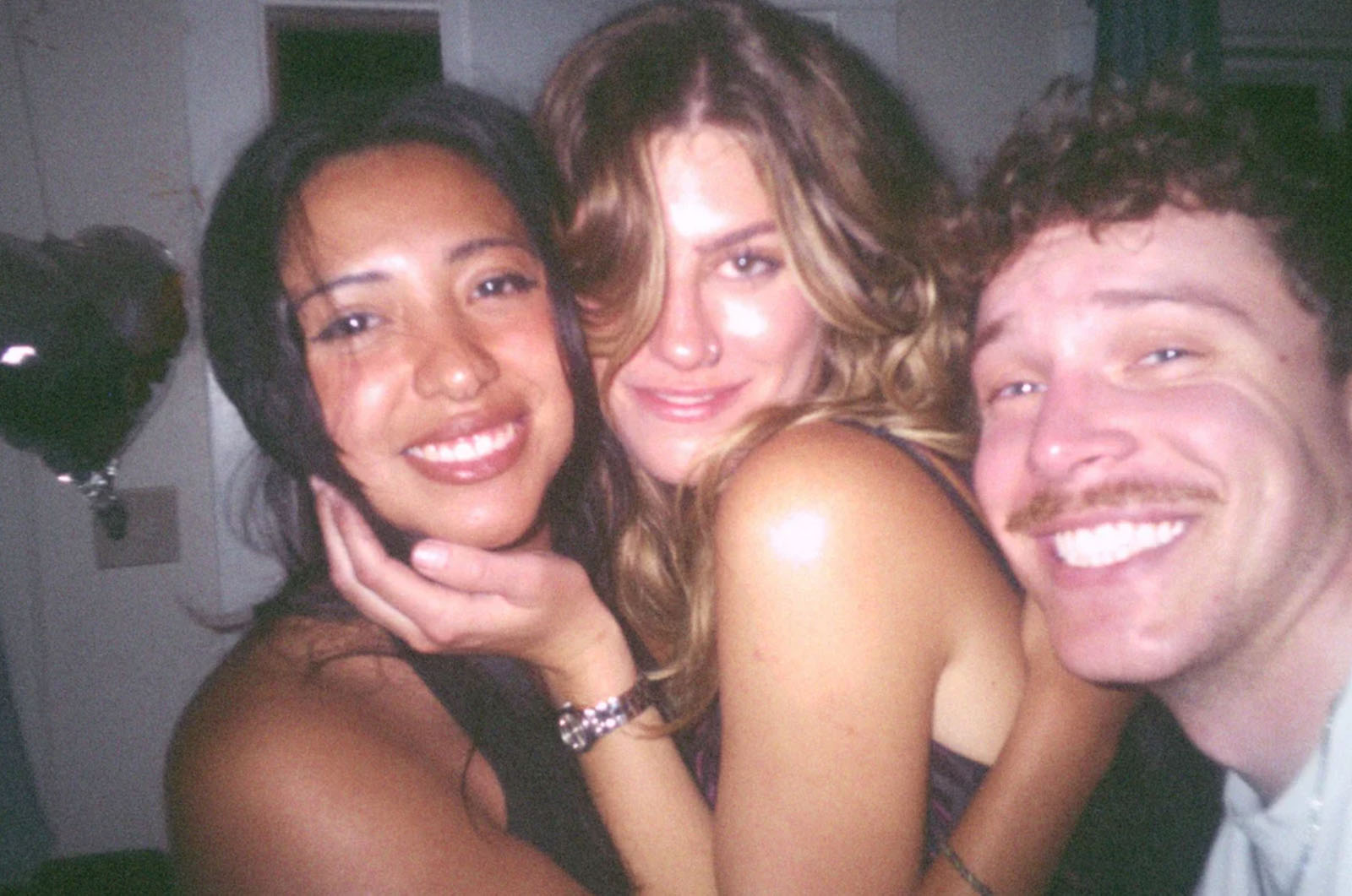 Three people smile closely together indoors, posing for a photo. Two women are in the center, one with dark hair and one with light hair, and a man with curly hair and a mustache is on the right. Dimly lit background.