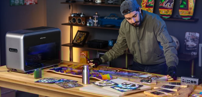 A man in a gray cap and jacket works on colorful art projects at a wooden table, using tools and a modern printer in a creative studio with decorated skateboards and shelves in the background.