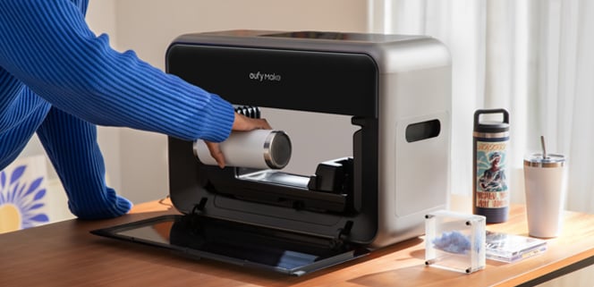 A person places a vacuum cylinder into a compact, modern-looking cleaning appliance on a wooden table, with drink containers and small objects nearby. Sunlight filters through a window in the background.