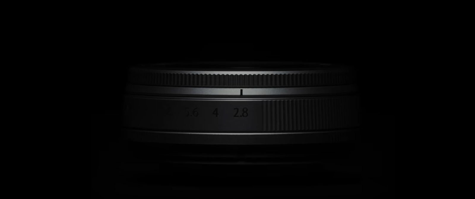 A camera lens is partially illuminated against a black background, with its ridged focus and zoom rings visible and aperture markings faintly seen.