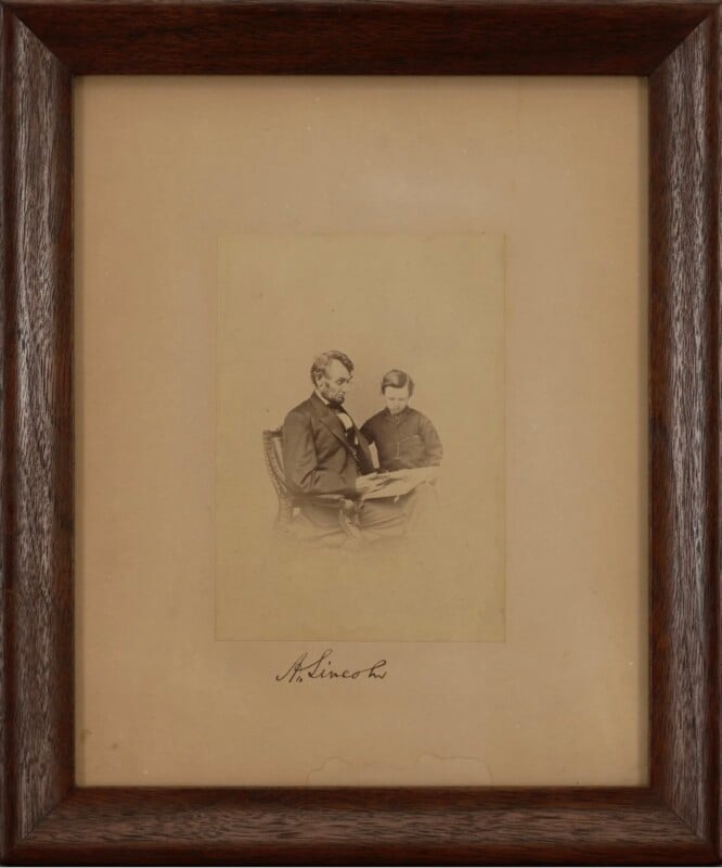 A sepia-toned photograph of Abraham Lincoln sitting in a chair, reading a book with a young boy beside him. The image is framed in dark wood, with “A. Lincoln” signed below the photograph.
