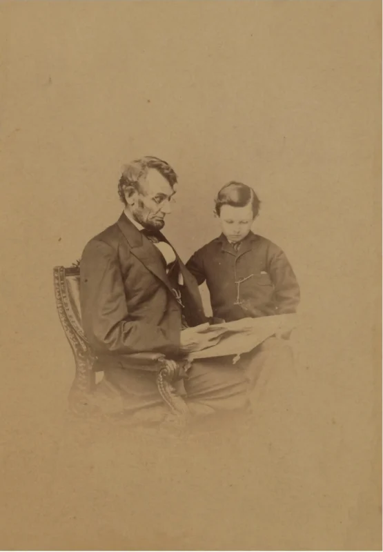 A sepia-toned photograph of Abraham Lincoln seated on a chair, reading a book with his young son, Tad, who stands at his side and looks at the pages. Both appear focused on the book.