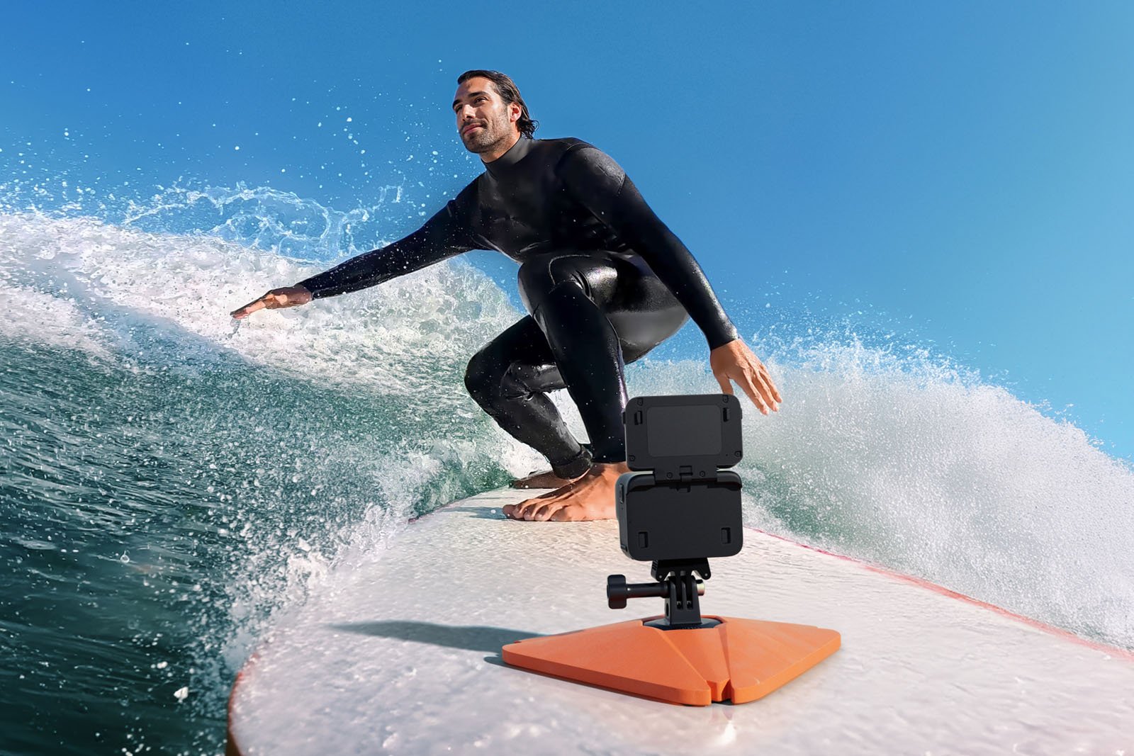 A surfer in a black wetsuit rides a wave on a white surfboard. A mounted action camera is attached to the front of the board, capturing the action against a clear blue sky.