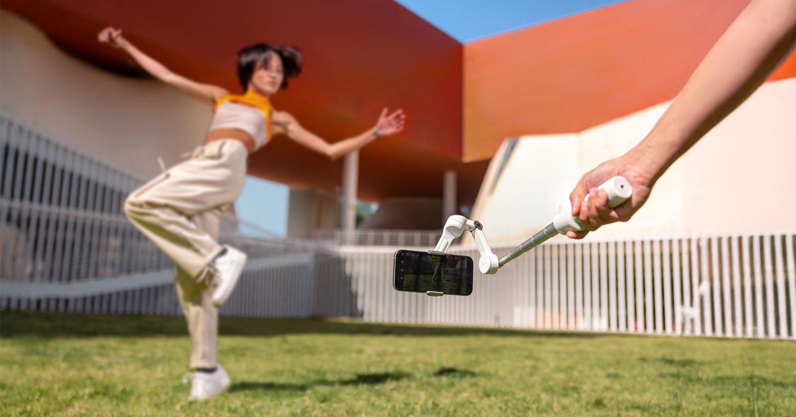 A person jumps joyfully on grass while another holds a selfie stick with a smartphone, capturing the moment. Modern buildings with white railings and red accents are in the background under a clear blue sky.