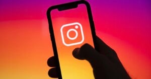 A hand holding a smartphone displaying the Instagram logo on the screen, with a vibrant pink, orange, and yellow gradient background.