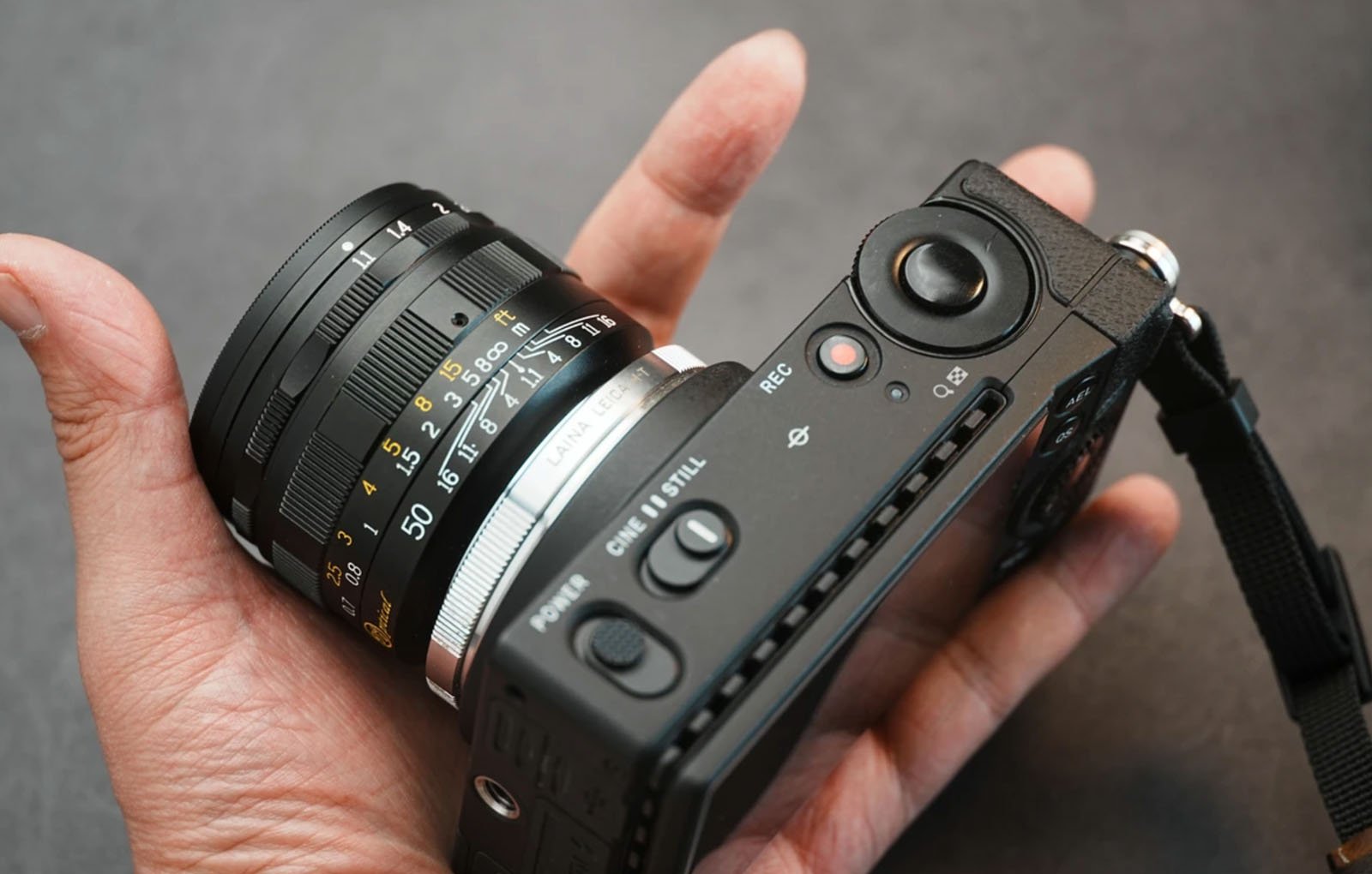 A hand holds a compact digital camera with a manual focus lens, showing dials, buttons, and lens markings on a dark background.