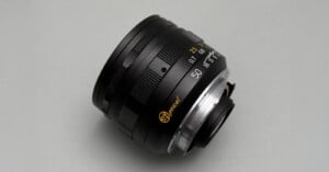 A close-up photo of a black camera lens with gold and white markings, including "50" for the focal length, set against a plain light gray background.