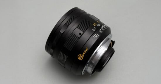 A close-up photo of a black camera lens with gold and white markings, including "50" for the focal length, set against a plain light gray background.