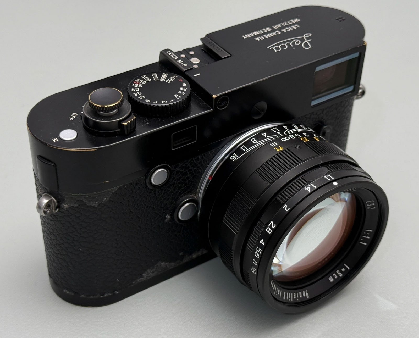 A black Leica camera with a textured grip, silver dials, and a large lens. The camera shows signs of wear, especially on the bottom edge. The Leica logo is visible on the top right.