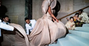 A person in an elaborate, flowing beige gown walks up stairs while attendants in white help with the dress. Several people in gold outfits sit on the steps, some eating and chatting, against a stone wall with engraved text.