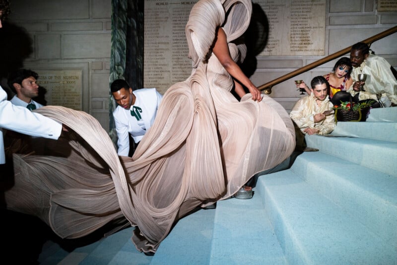 A person in an elaborate, flowing gown ascends blue steps while others in formal attire assist and watch, some seated on the stairs, against a backdrop of engraved stone walls.