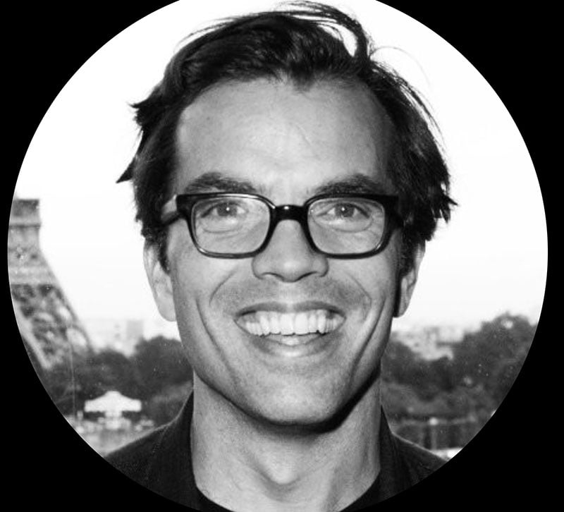 A man with dark hair and glasses smiles broadly at the camera. The photo is black and white, with a blurred outdoor background and part of a metal structure visible on the left.