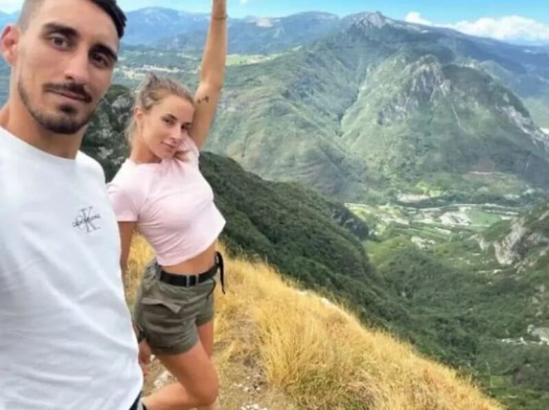 A man and a woman pose for a selfie on a grassy mountain edge with green valleys and distant mountains in the background, under a partly cloudy sky. The woman raises one arm and both appear happy.