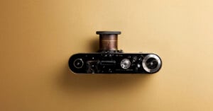 A vintage black and silver camera with a protruding brass lens is placed on a plain, light brown background, viewed from above.