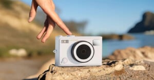 A hand gently touches a white Lenovo camera resting on a rock outdoors, with a blurred natural landscape and blue sky in the background.