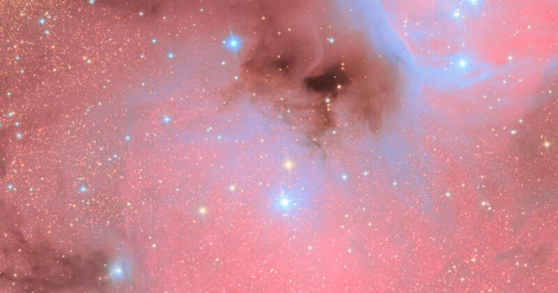 A vibrant view of outer space shows countless stars scattered across a pink and blue nebula, with dark dust clouds creating swirling patterns in the colorful cosmic clouds.
