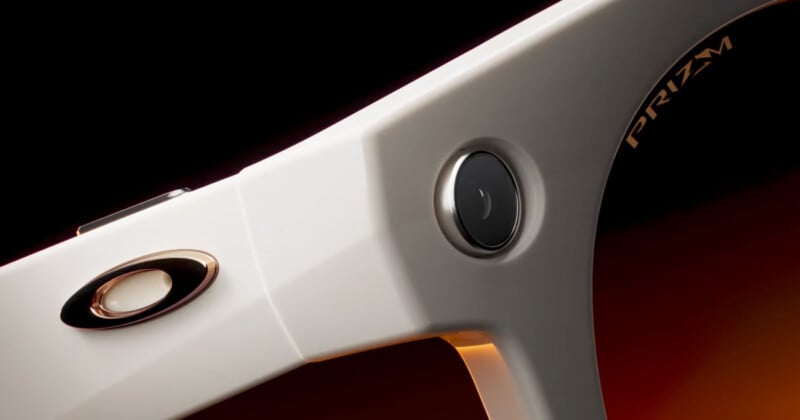 Close-up of the side of white Oakley sunglasses featuring a small built-in camera lens and the "Prizm" logo on the lens, set against a gradient orange-black background.