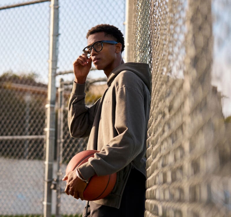 A young man wearing glasses and a hoodie stands by a chain-link fence on a basketball court, holding a basketball under one arm and adjusting his glasses with his other hand.