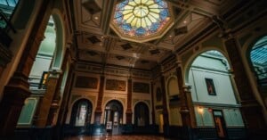 A grand hall with high ceilings, ornate columns, arched doorways, and a colorful stained glass skylight, illuminated by natural light streaming in. The architecture is classical and elegant.
