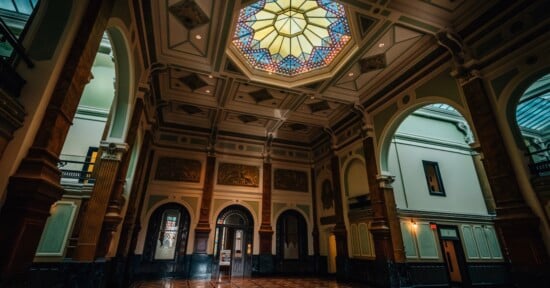 A grand hall with high ceilings, ornate columns, arched doorways, and a colorful stained glass skylight, illuminated by natural light streaming in. The architecture is classical and elegant.