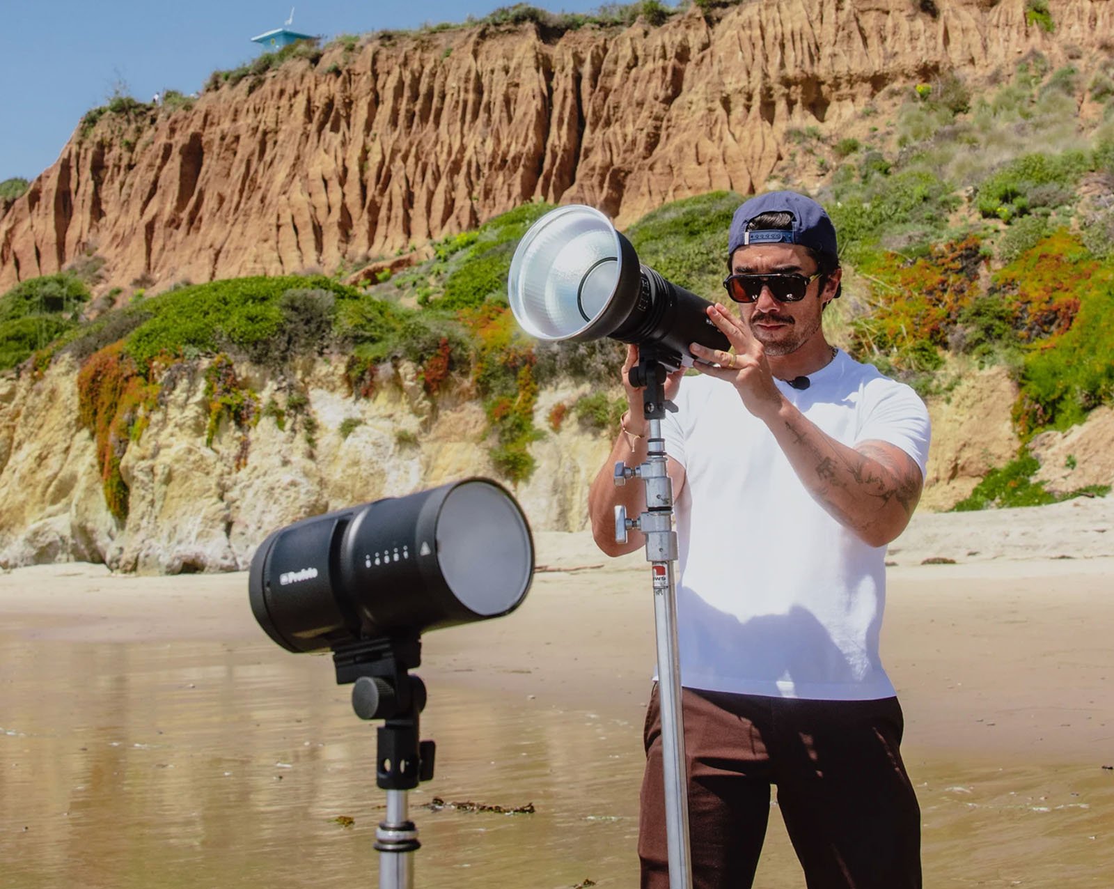 A man in sunglasses and a white shirt adjusts studio lighting equipment on a sandy beach, with rocky cliffs and greenery in the background.