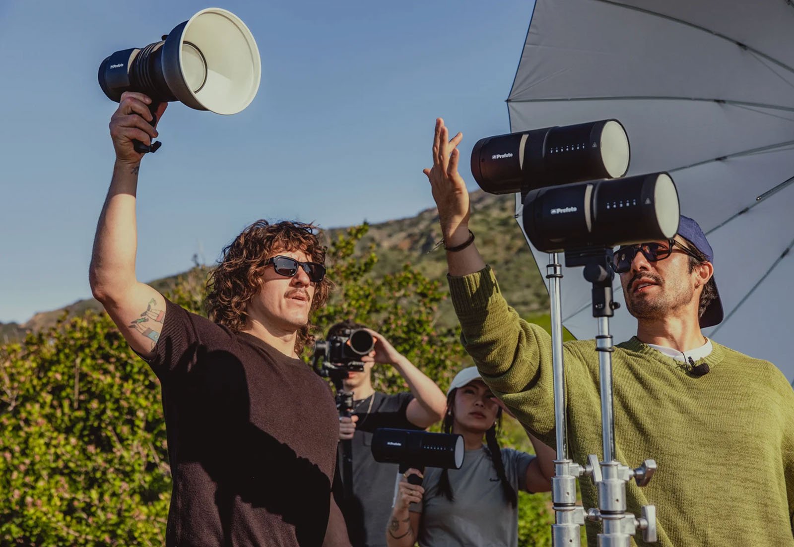 Two people wearing sunglasses adjust professional lighting equipment outdoors, with a third person filming and hills in the background. A large white umbrella reflector is visible nearby.