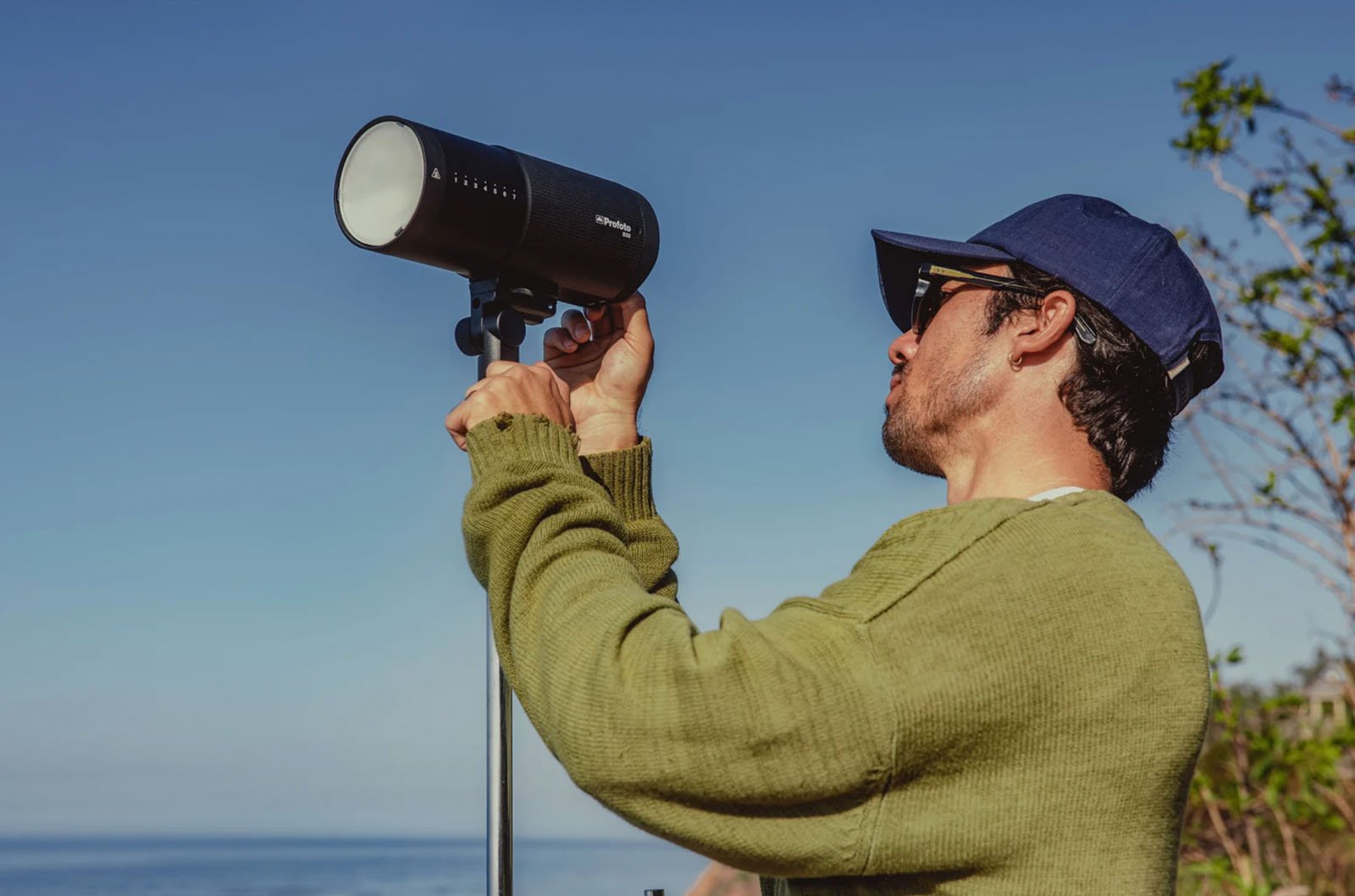 A person wearing a green sweater, blue cap, and sunglasses adjusts a black telescope or camera on a tripod, outdoors near the water under a clear blue sky.