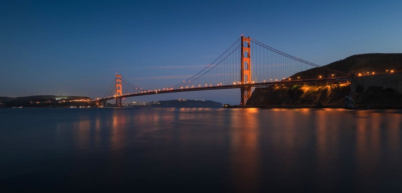 Golden Gate Bridge illuminated at dusk, stretching over calm water with city lights in the distance and hills silhouetted against a deep blue sky.