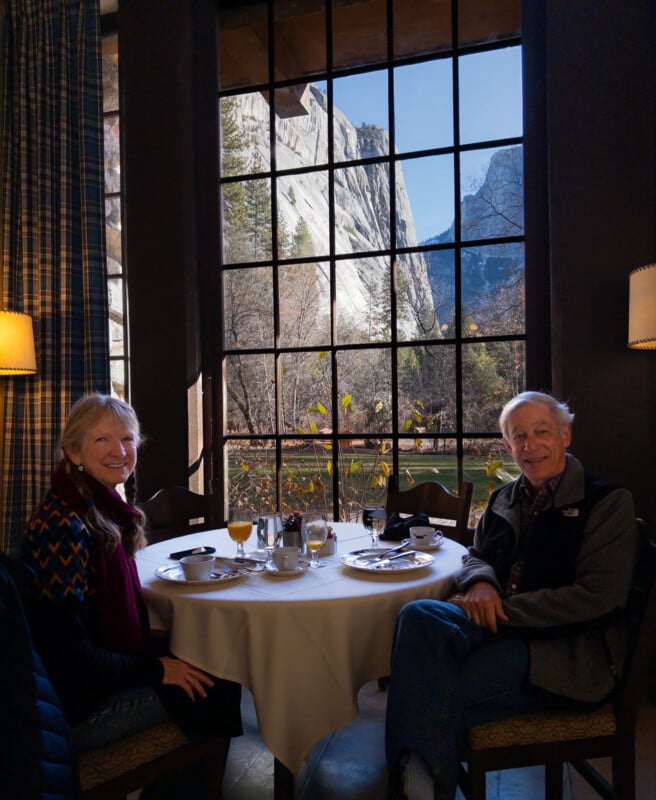 An older woman and man sit smiling at a round table set for a meal, in front of a large window with a scenic view of mountains, trees, and a blue sky in the background.