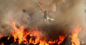 A person stands amid thick smoke, raising a Palestinian flag above intense flames and burning debris, creating a dramatic and chaotic scene.