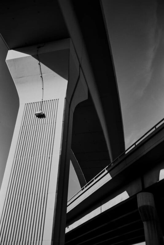Black and white photo of a tall highway overpass and its thick supporting pillars, viewed from below, with dramatic shadows and upward perspective emphasizing the structure’s size and geometric lines.