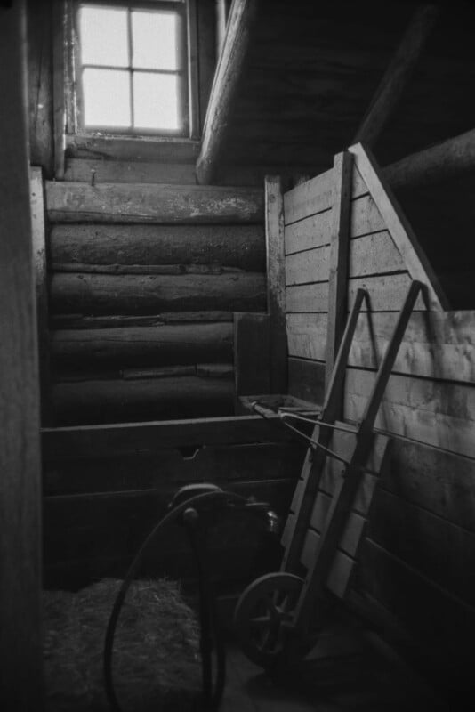 Black and white photo of a rustic wooden barn interior with a handcart leaning against a wooden wall, a pile of hay, and a window above wooden steps letting in light.