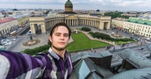 A young man in a plaid shirt takes a selfie from a high vantage point, with the Kazan Cathedral and a cityscape visible in the background on a cloudy day.