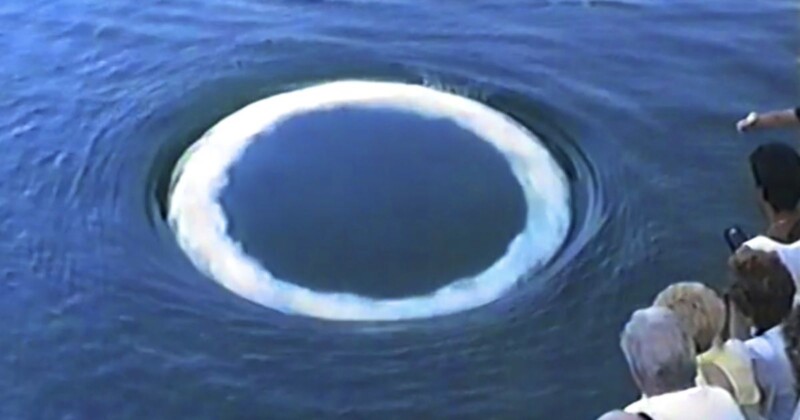 A group of people look at a large, circular whirlpool with a white frothy rim forming in the blue water.