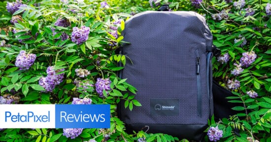 A black, grid-patterned Shimoda backpack sits among lush green leaves and purple flowers. Overlay text reads "PetaPixel Reviews" in the lower left corner.
