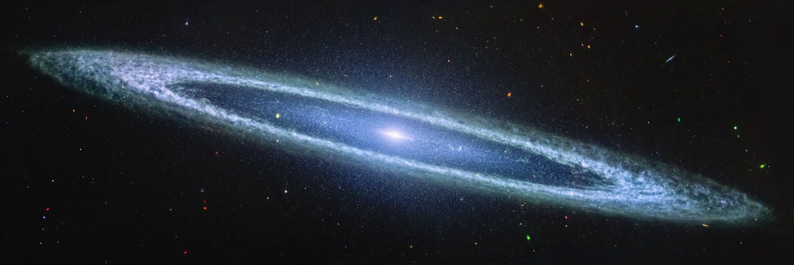 A bright, spiral galaxy is seen edge-on, glowing with a luminous center and long, thin arms. Surrounding it are scattered stars and distant galaxies in the dark expanse of space.