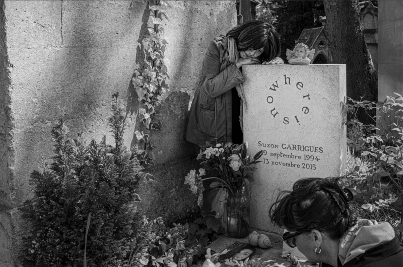 A woman leans in grief on a gravestone that reads "nowhere, now here, us!" with flowers and photos at its base; another woman kneels nearby, facing the grave. The scene is in black and white.