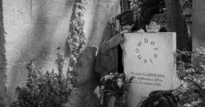 A woman leans on a gravestone, appearing to grieve. The gravestone reads "Nowhere! Us!" and marks the grave of Suzon Garrigues (1994–2015). Flowers and a small angel statue are placed in front of the stone.