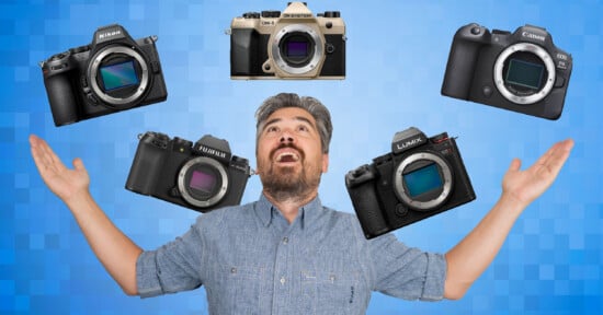 A man with a beard and gray hair stands with arms raised, looking upward, as five mirrorless cameras from different brands float above him against a blue, checkered background.