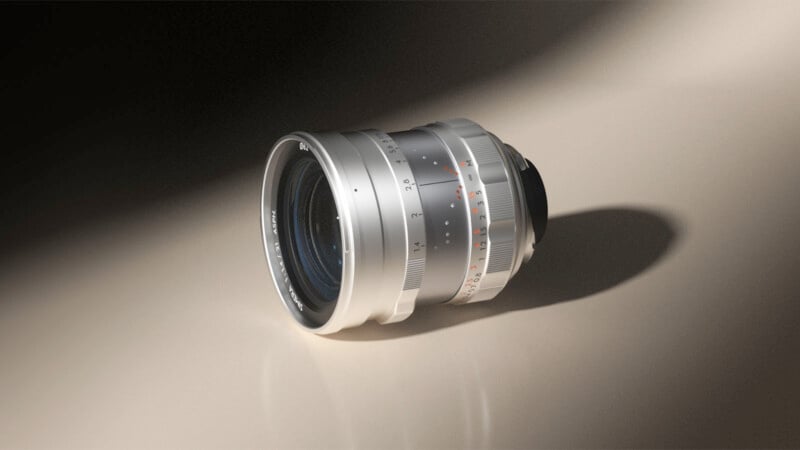 A silver camera lens with manual focus and aperture markings rests on a smooth, light-colored surface, casting a soft shadow to the right.