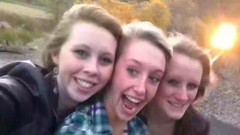 Three young women smiling and posing closely together for a selfie outdoors, with autumn trees and sunlight in the background.