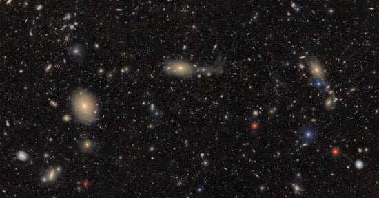 A deep space image showing countless galaxies and stars scattered across a dark background, with some galaxies appearing as bright, fuzzy spots and others as colorful points of light in blue, red, and yellow hues.