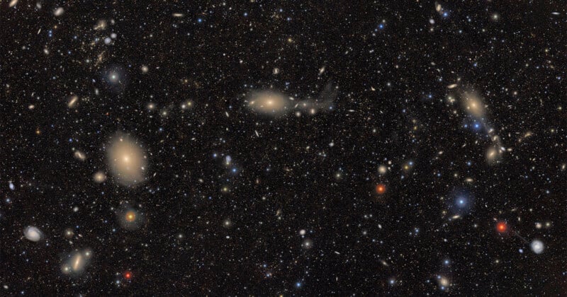 A deep space image showing countless galaxies and stars scattered across a dark background, with some galaxies appearing as bright, fuzzy spots and others as colorful points of light in blue, red, and yellow hues.