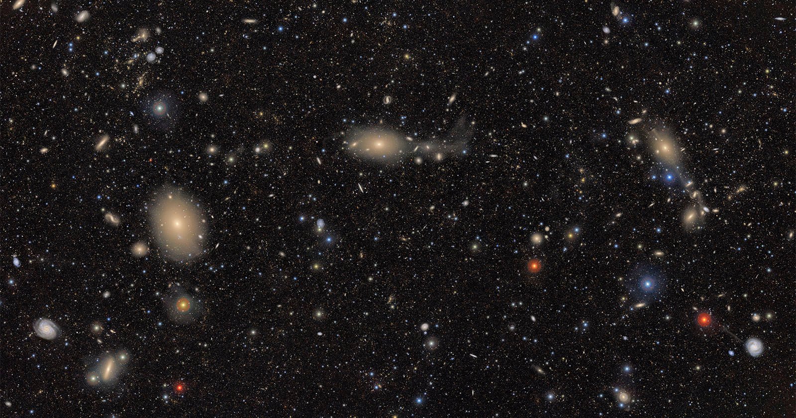 A deep space image showing countless galaxies and stars scattered across a dark background, with some galaxies appearing as bright, fuzzy spots and others as colorful points of light in blue, red, and yellow hues.